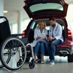 Folding travel power wheelchair by a car trunk and airport area with clinician and caregiver reviewing Medicare and insurance paperwork